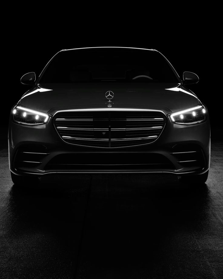 Front view of a dark Mercedes-Benz with its headlights on against a black background.