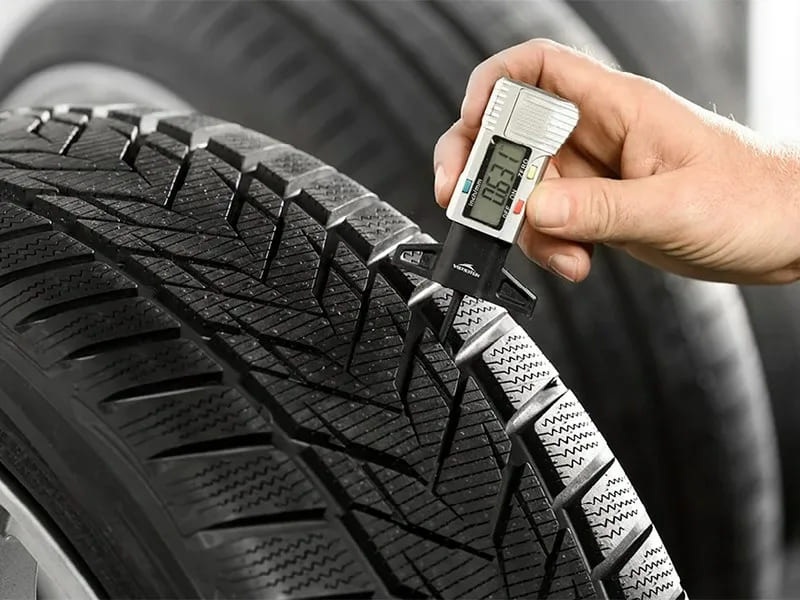 A hand measures the tread depth of a car tyre with a digital gauge.