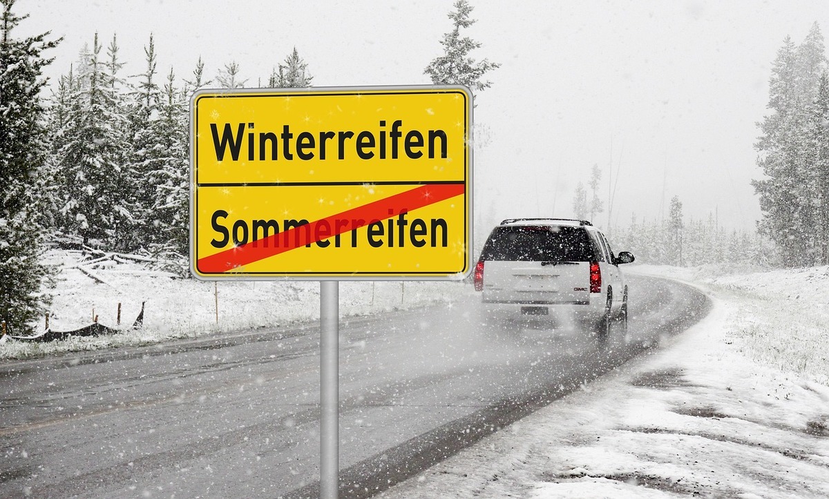 Snow-covered road with a car and a yellow sign reading “Winterreifen”, with “Sommerreifen” crossed out.
